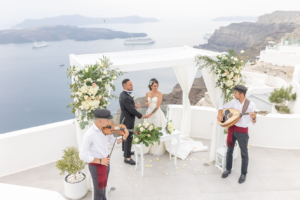 Cérémonie de mariage à Santorin : les mariés et deux musiciens sur une terrasse blanche au-dessus de la caldeira